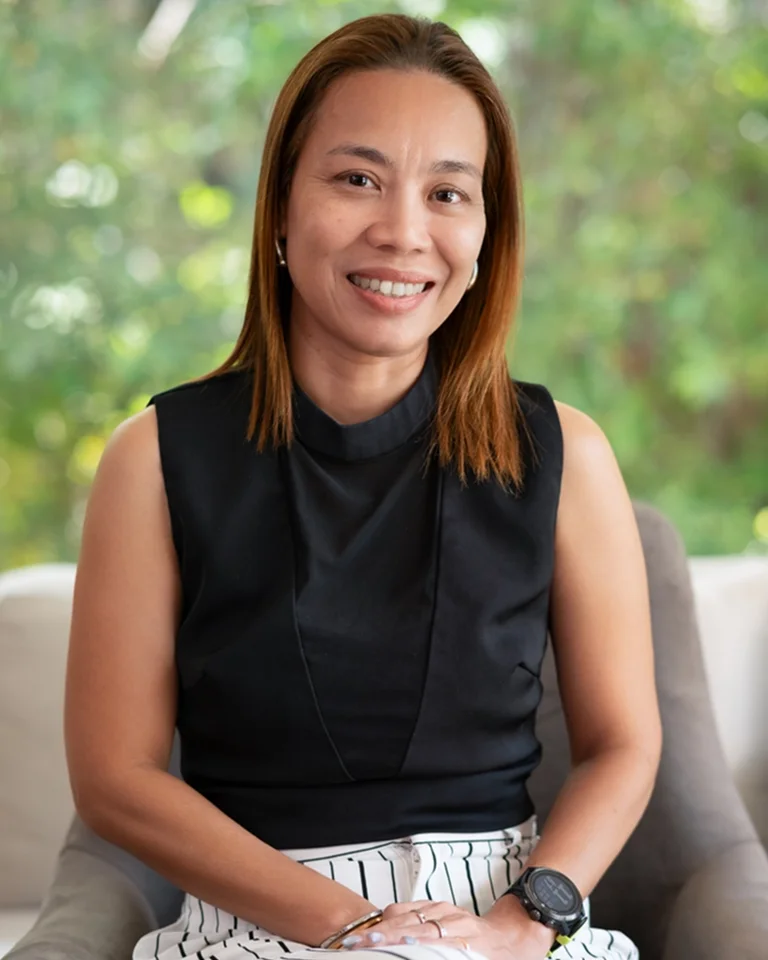 Smiling woman seated on a couch outdoors in front of greenery, wearing a black sleeveless top and striped white pants with a wristwatch.