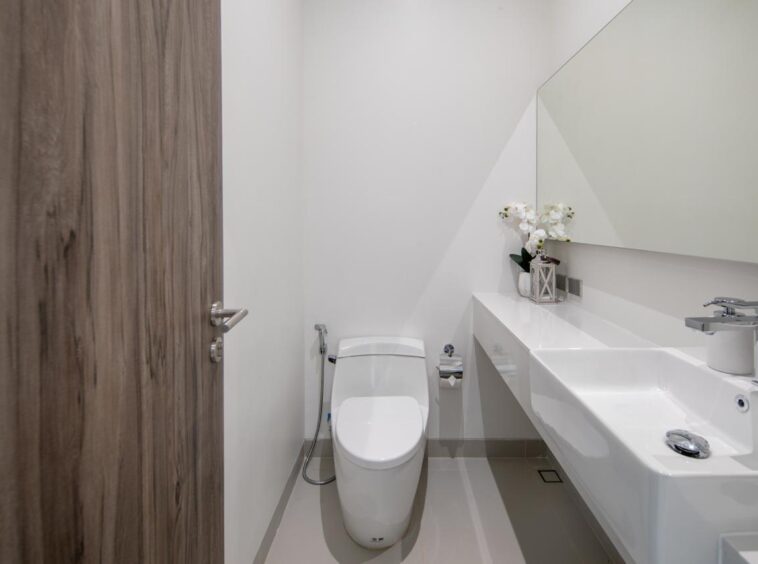 Small modern bathroom with a white toilet and a long white countertop sink, mirror, and flower vase.