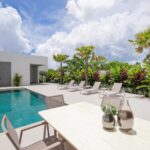 Modern poolside patio with a rectangular pool, white concrete deck, lounge chairs, and tropical plants along a privacy hedge.