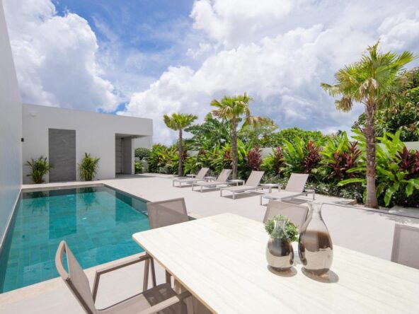 Modern poolside patio with a rectangular pool, white concrete deck, lounge chairs, and tropical plants along a privacy hedge.