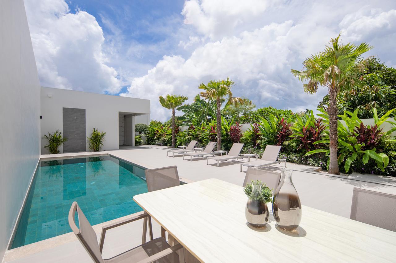 Modern poolside patio with a rectangular pool, white concrete deck, lounge chairs, and tropical plants along a privacy hedge.