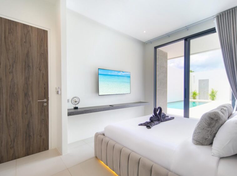Minimalist hotel bedroom with a large bed, gray throw pillows, and sliding glass doors to a poolside view outside home-like balcony area.