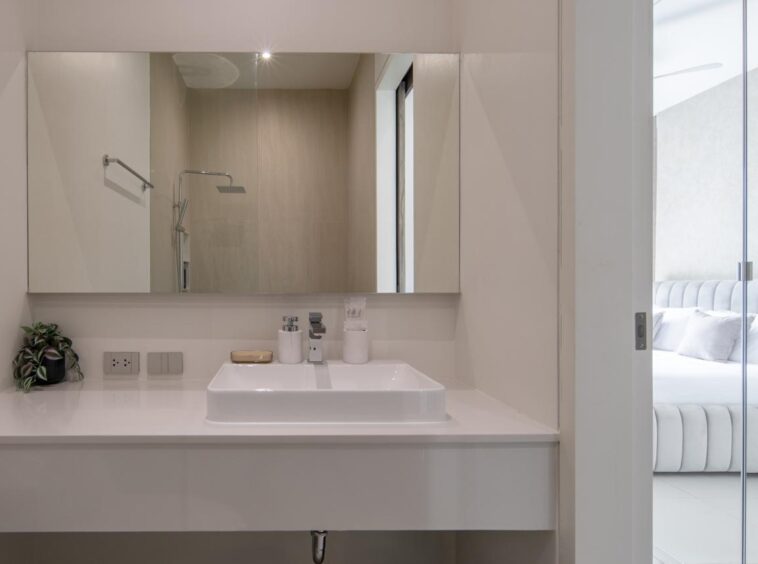 Modern white bathroom vanity with square sink, chrome faucet, and small plant; glass-walled bedroom visible to the right.