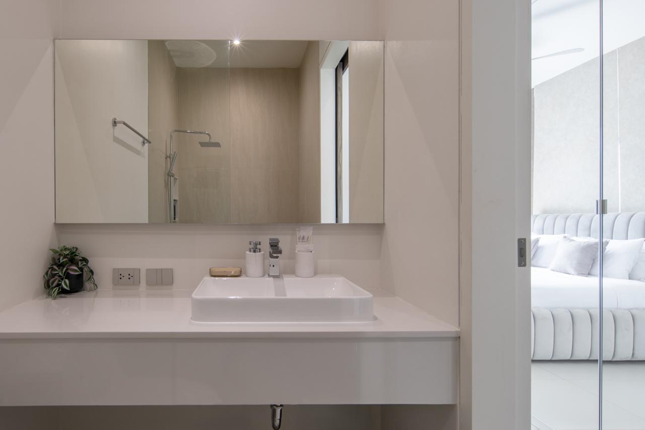 Modern white bathroom vanity with square sink, chrome faucet, and small plant; glass-walled bedroom visible to the right.