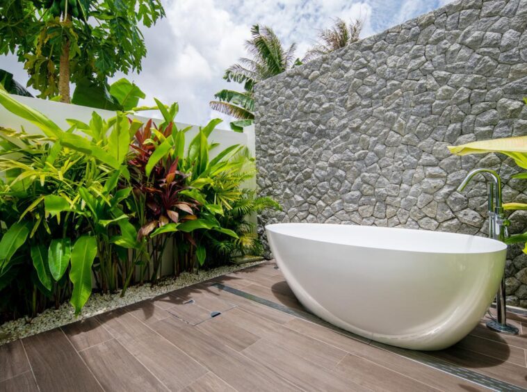 Outdoor freestanding white bathtub on a wooden deck beside a textured stone wall and tropical plants.