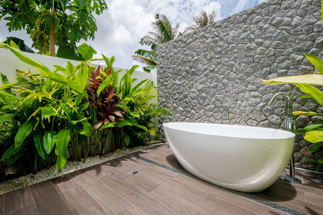 Outdoor freestanding white bathtub on a wooden deck beside a textured stone wall and tropical plants.