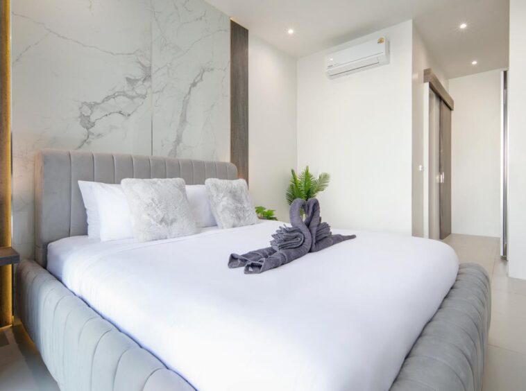 Luxurious hotel bedroom with a gray upholstered bed, white linens, marble accent wall, and a towel swan centerpiece on the duvet