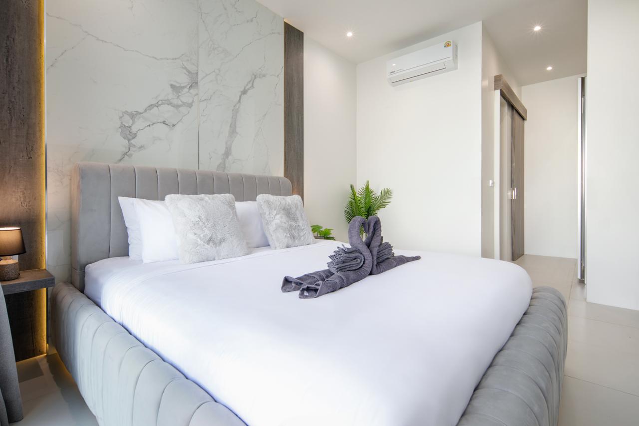 Luxurious hotel bedroom with a gray upholstered bed, white linens, marble accent wall, and a towel swan centerpiece on the duvet