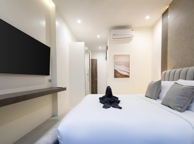 Bright hotel bedroom with a wall-mounted flat-screen TV, a large upholstered headboard, white bedding, and gray accent pillows; a folded towel centerpiece sits on the bed.