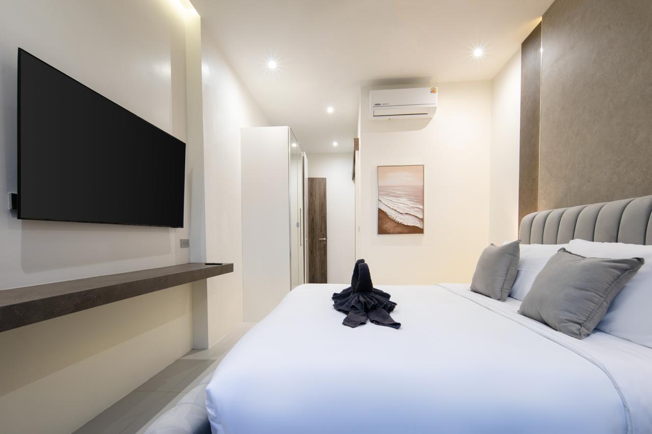 Bright hotel bedroom with a wall-mounted flat-screen TV, a large upholstered headboard, white bedding, and gray accent pillows; a folded towel centerpiece sits on the bed.