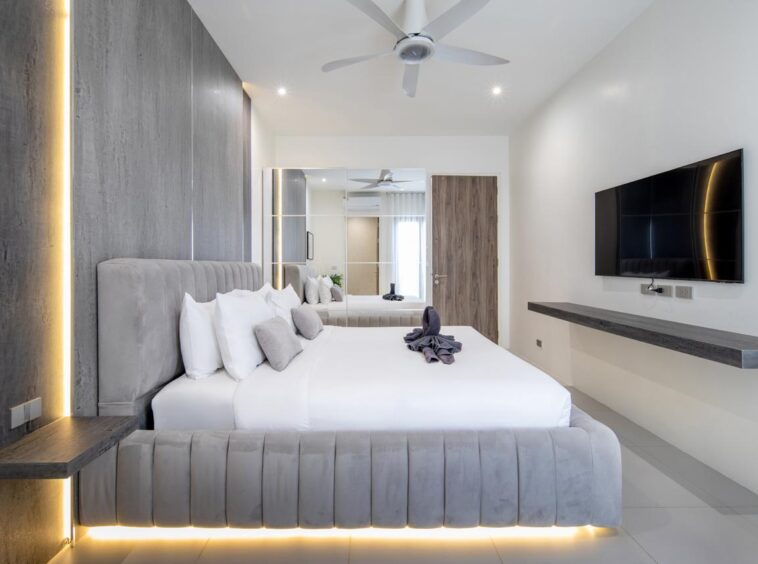 Modern hotel bedroom with a large bed, grey tufted headboard, white linens, and warm LED lighting along the base; wall-mounted TV and floating desk visible on the right.
