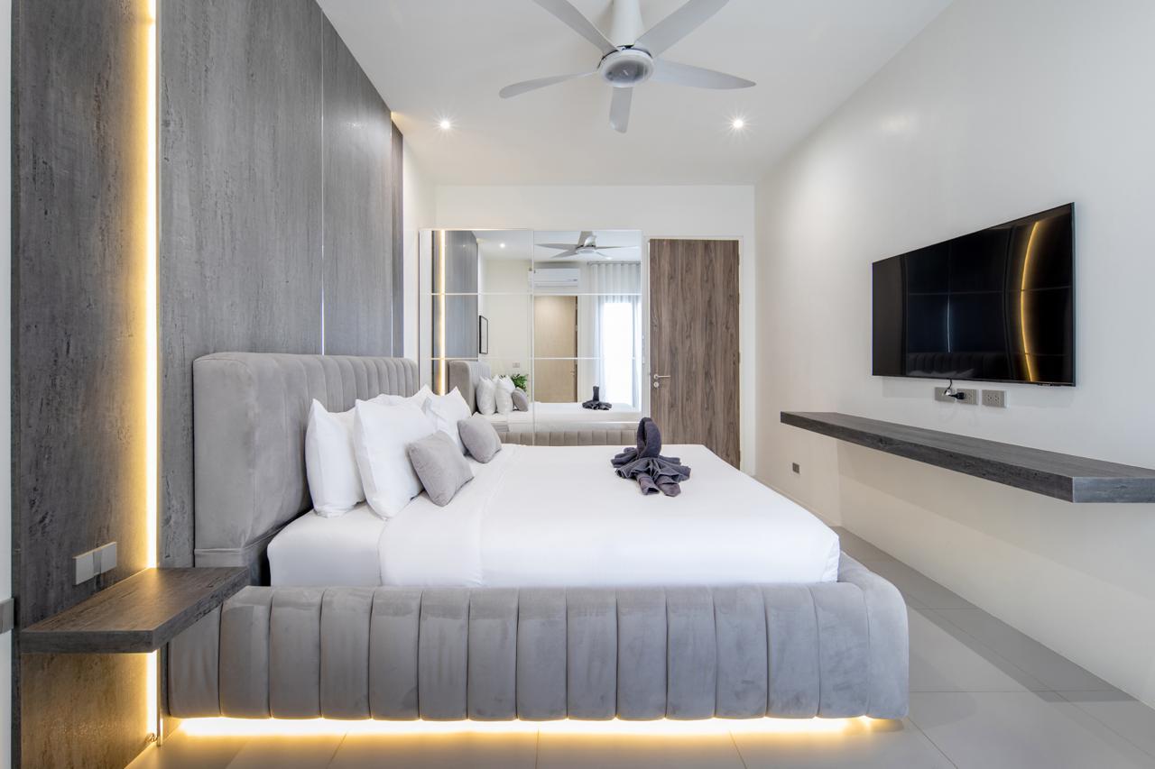 Modern hotel bedroom with a large bed, grey tufted headboard, white linens, and warm LED lighting along the base; wall-mounted TV and floating desk visible on the right.