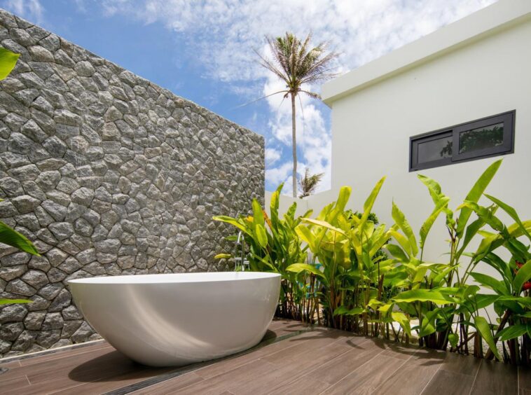 White freestanding bathtub on a wooden deck beside a textured stone wall and lush tropical plants, with a modern white building and blue sky above.