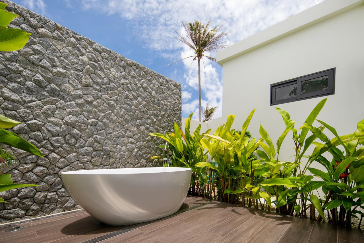 White freestanding bathtub on a wooden deck beside a textured stone wall and lush tropical plants, with a modern white building and blue sky above.
