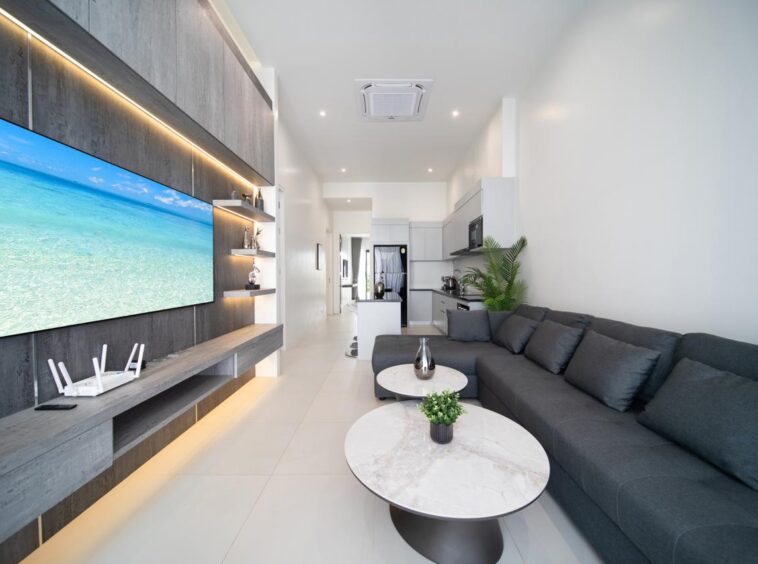 Open-concept living area with a dark gray sectional, two round marble coffee tables, and a wall-mounted jungle beach TV wall; kitchen in the background.