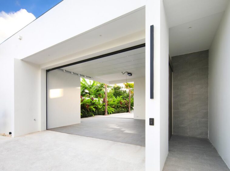 Modern white house with a wide open garage showing a courtyard and tropical plants beyond, and a grey tiled entry on the right side to a narrow hallway.