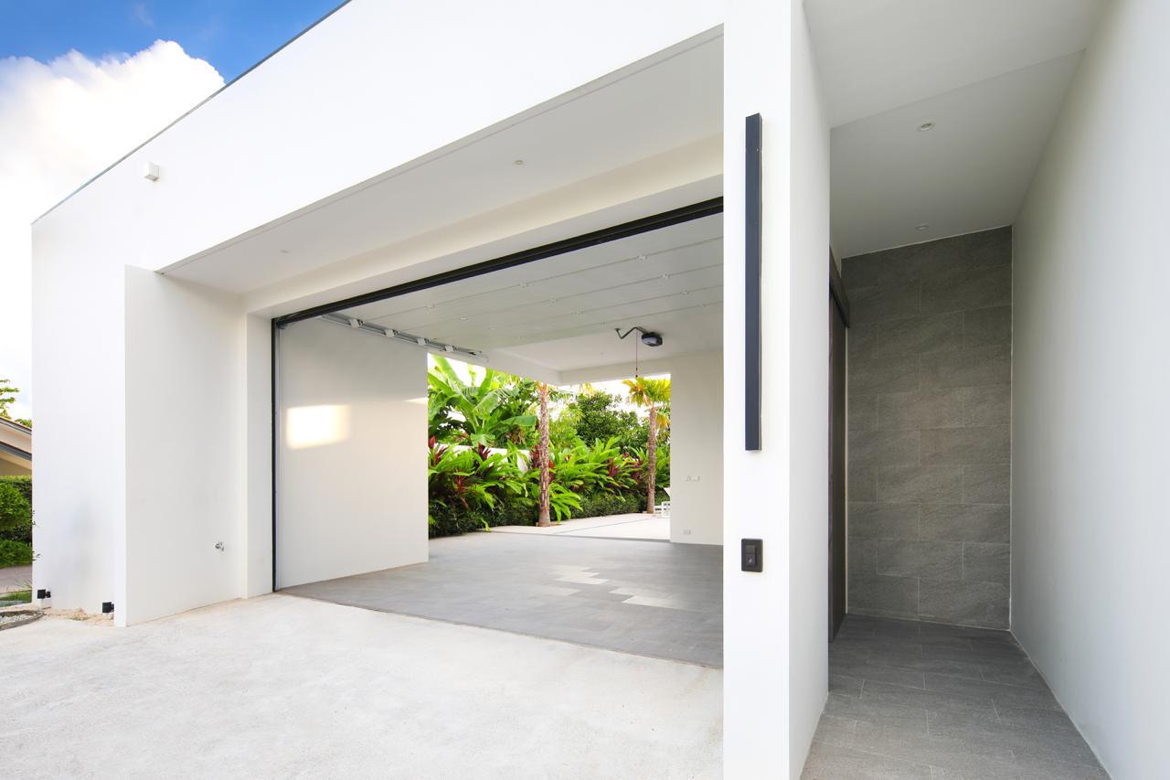 Modern white house with a wide open garage showing a courtyard and tropical plants beyond, and a grey tiled entry on the right side to a narrow hallway.