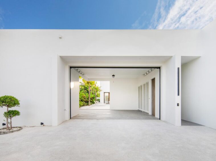 White modern house with a large open garage, concrete driveway, and a small trimmed tree on the left against a bright blue sky.
