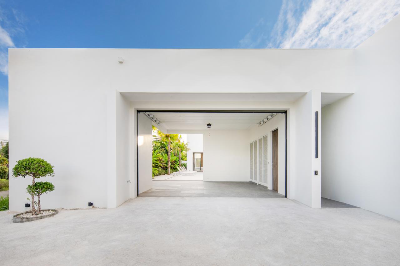 White modern house with a large open garage, concrete driveway, and a small trimmed tree on the left against a bright blue sky.
