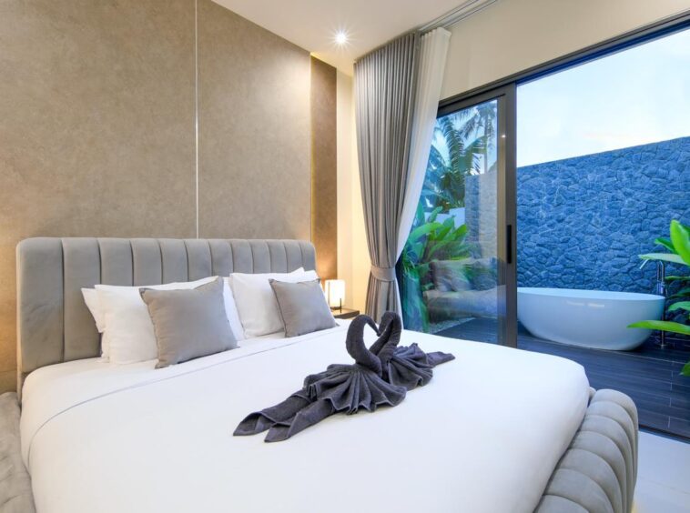 Luxurious hotel bedroom with a plush padded headboard, white linens, and gray pillows; glass door to an outdoor bathtub and greenery outside.