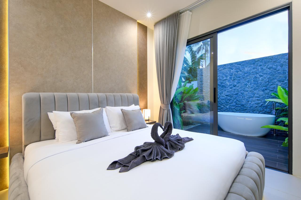Luxurious hotel bedroom with a plush padded headboard, white linens, and gray pillows; glass door to an outdoor bathtub and greenery outside.