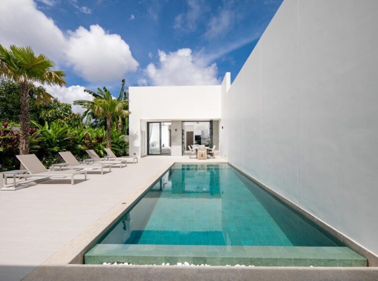 Modern white villa with a long rectangular pool, lounge chairs, and tropical plants under a bright blue sky.