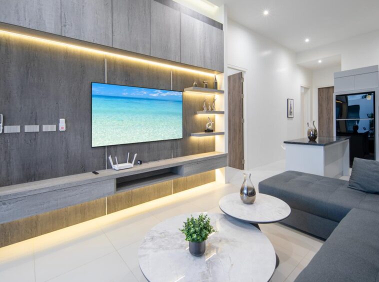 Contemporary living room with a wall-mounted TV on gray wood paneling, illuminated shelves, and a gray sectional sofa.