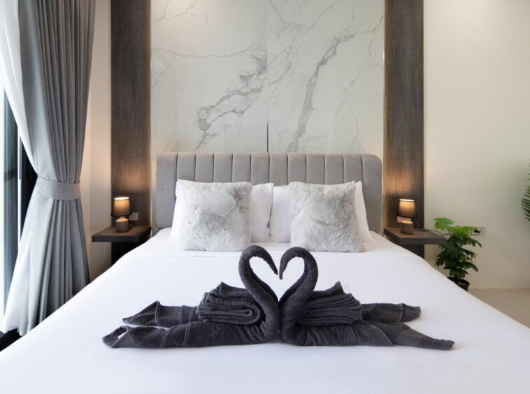 Luxurious bedroom with a tufted gray headboard, marble wall panels, and swan-shaped towels on a white duvet.