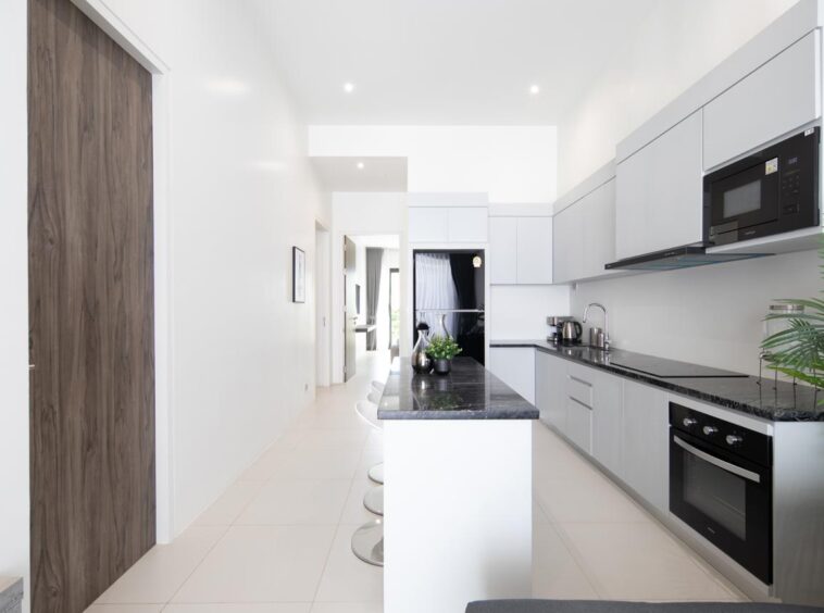 Bright modern kitchen with white walls, grey cabinets, black countertops, and stainless-steel appliances along a long counter.