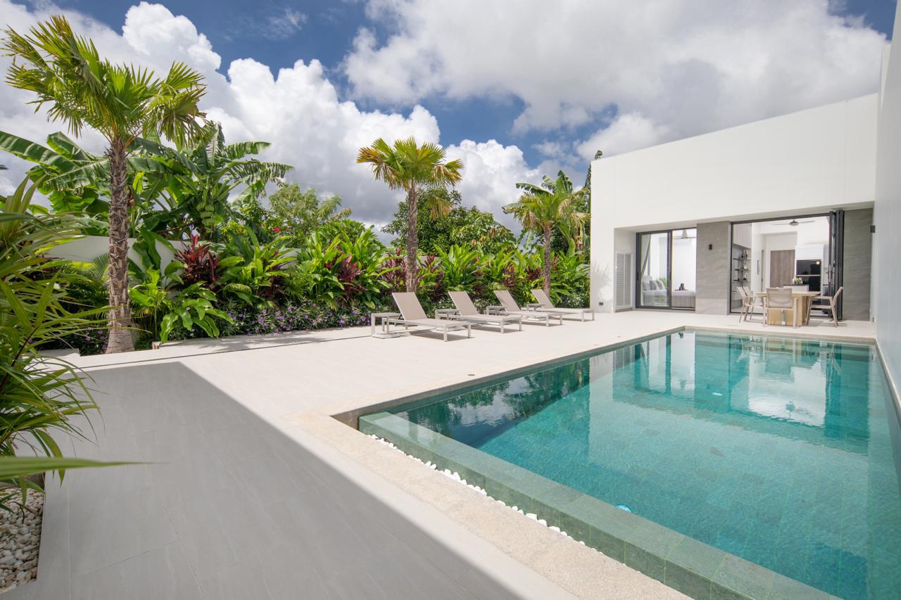Modern white villa with a rectangular pool, beige lounge chairs, tropical plants, and an open living area.
