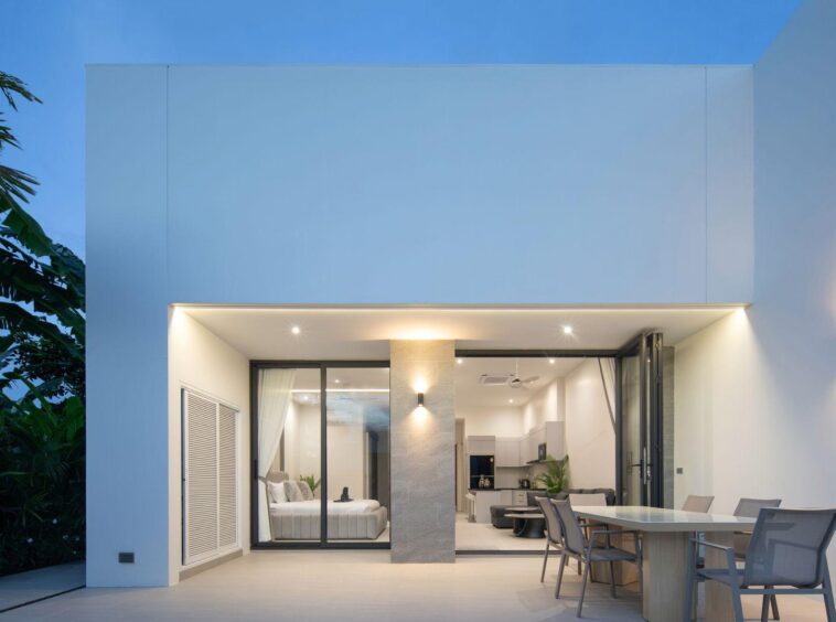 Modern white house with large glass sliding doors opening to a lit interior: a bedroom to left and an open-plan kitchen/living area, outdoor dining table on patio.