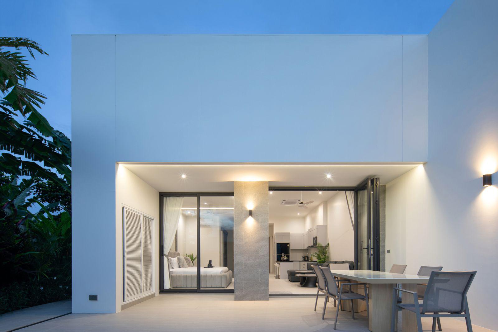 Modern white house with large glass sliding doors opening to a lit interior: a bedroom to left and an open-plan kitchen/living area, outdoor dining table on patio.