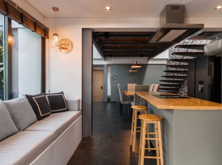 Open-plan kitchen and living area with a wooden island breakfast bar and three stools, gray seating along a large window on the left, and a floating staircase in the background.