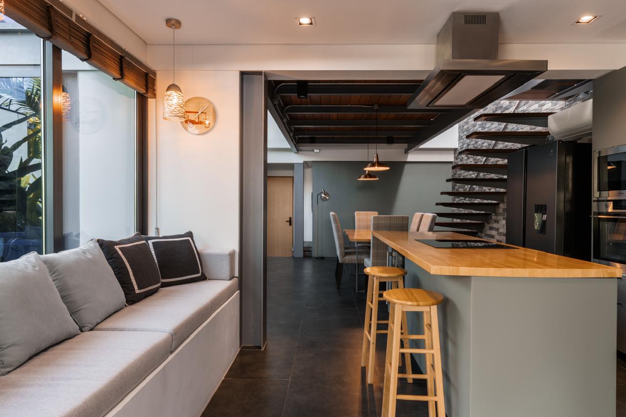 Open-plan kitchen and living area with a wooden island breakfast bar and three stools, gray seating along a large window on the left, and a floating staircase in the background.