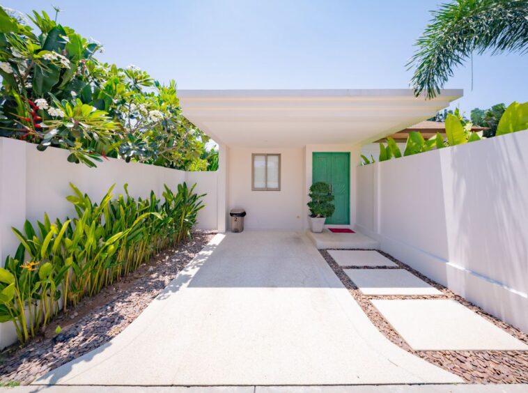 Bright, modern home entry with a green door and white walls, flanked by tropical plants and a concrete walkway.