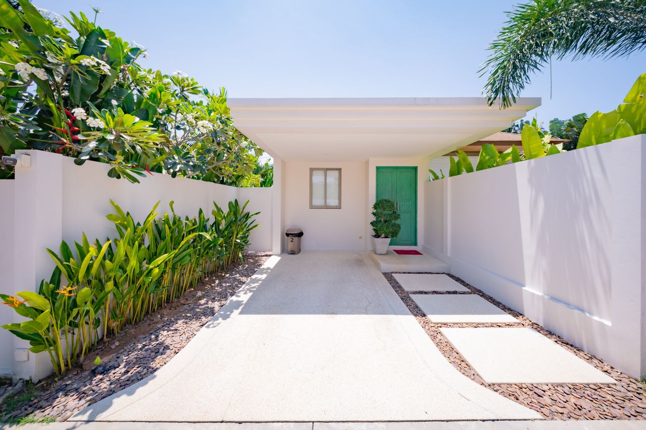 Bright, modern home entry with a green door and white walls, flanked by tropical plants and a concrete walkway.