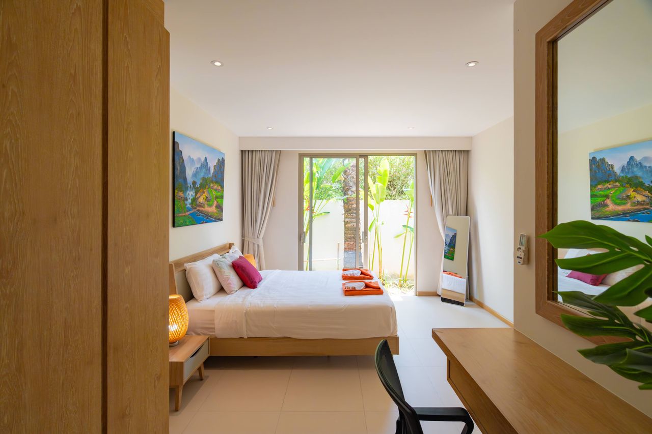 Bright hotel bedroom with a king bed, wooden furniture, and sliding glass doors to a tropical garden outside the room