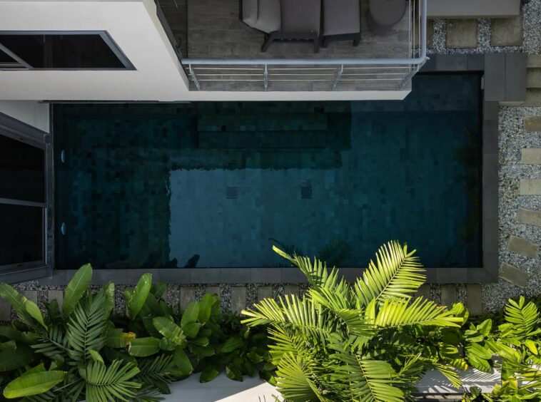 Aerial view of a rectangular swimming pool bordered by a stone path, with tropical plants in the foreground and a white building with a balcony.