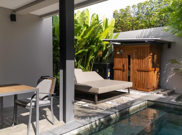 Outdoor patio with a wooden dining table and four chairs, a beige cushioned lounge bed, and a small pool nearby amidst tropical plants and a wooden shed in the background.
