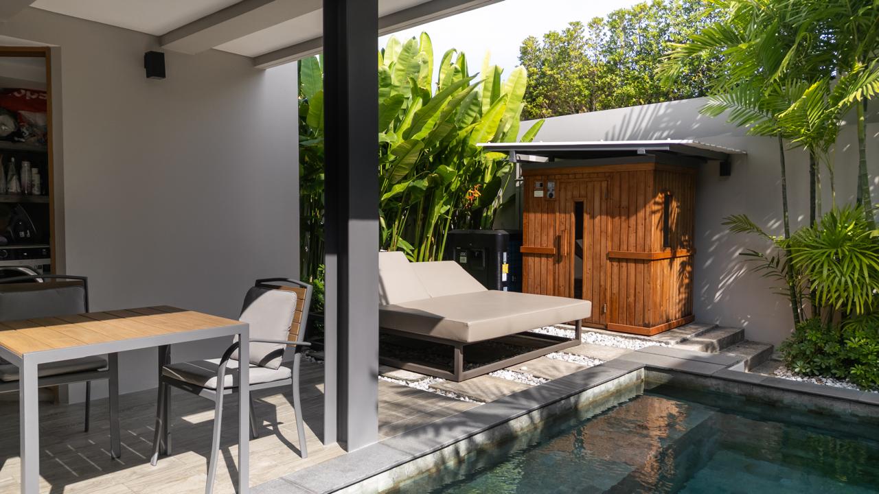 Outdoor patio with a wooden dining table and four chairs, a beige cushioned lounge bed, and a small pool nearby amidst tropical plants and a wooden shed in the background.