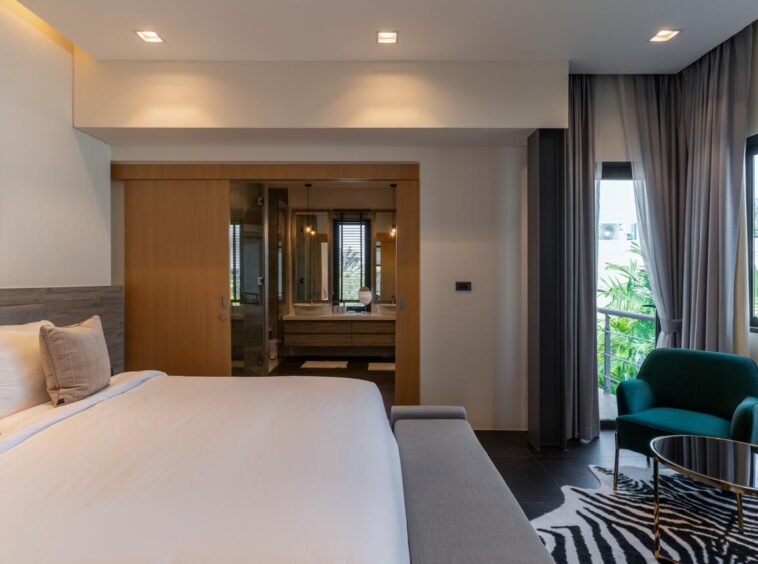 Hotel bedroom with a large white bed, gray headboard, and a bench at the foot, next to a seating area by the window.