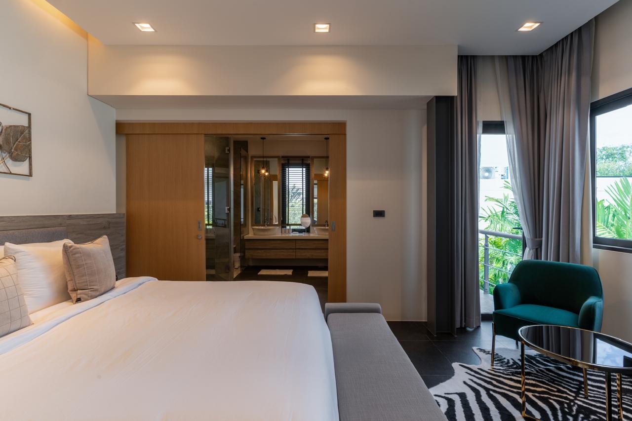 Hotel bedroom with a large white bed, gray headboard, and a bench at the foot, next to a seating area by the window.