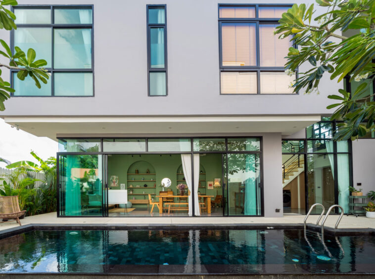 Modern two-story home with large glass sliding doors, pool in foreground, and tropical plants on sides.