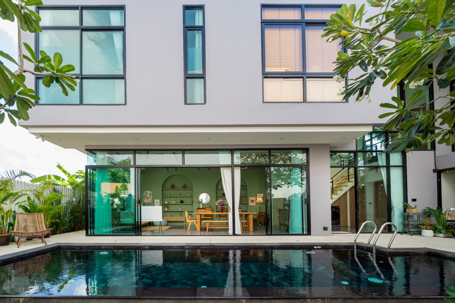 Modern two-story home with large glass sliding doors, pool in foreground, and tropical plants on sides.