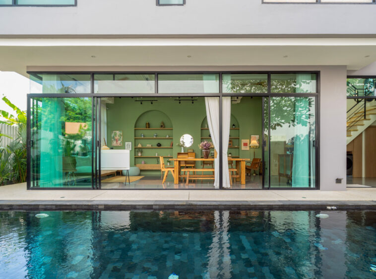 Open-plan living room with a green wall and wooden dining table seen through large glass sliding doors by a poolside patio.