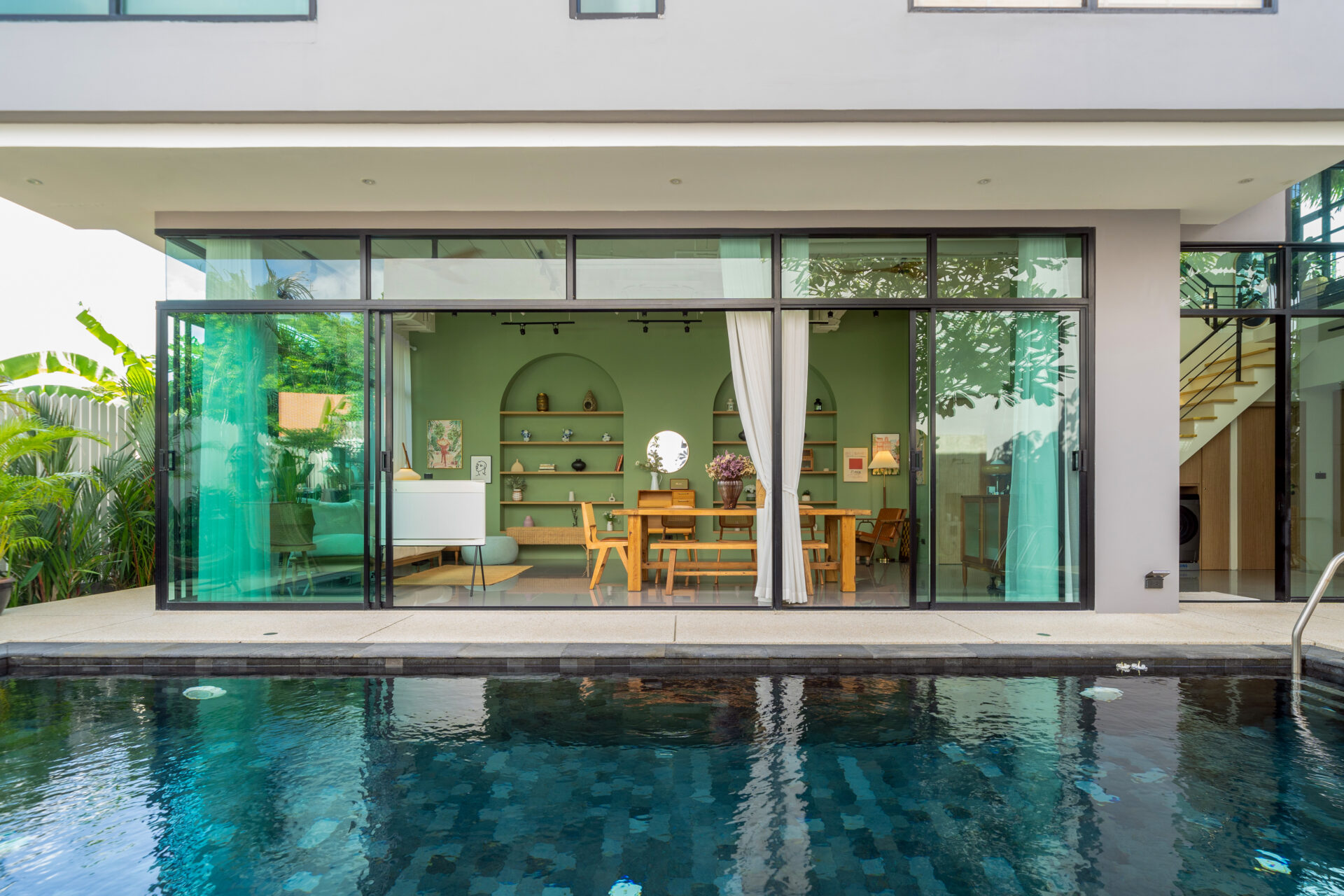 Open-plan living room with a green wall and wooden dining table seen through large glass sliding doors by a poolside patio.