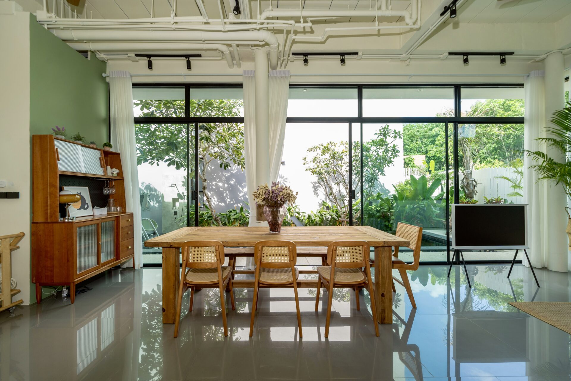 Open-plan dining room with a wooden table and chairs, large glass doors showing a lush garden outside.