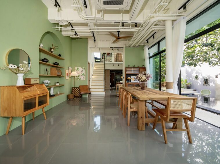 Open-plan dining and living area with green walls, a long wooden table and chairs, and large glass doors to an outdoor garden.