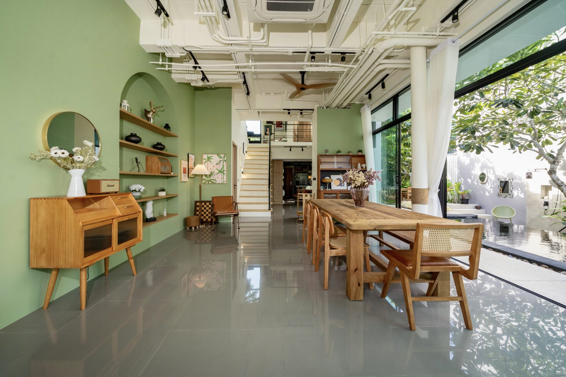 Open-plan dining and living area with green walls, a long wooden table and chairs, and large glass doors to an outdoor garden.