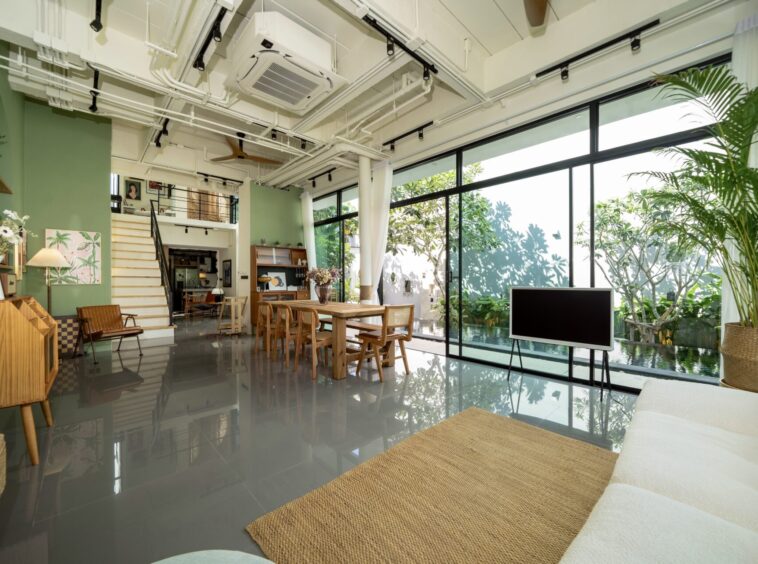 Open-concept living/dining area with large glass walls, wooden furniture, and lush indoor plants by a garden view.